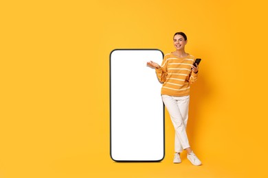 Image of Beautiful young woman with smartphone standing near big mobile phone with blank screen on golden background