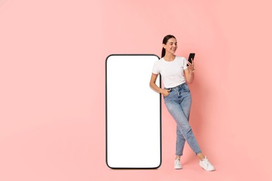 Beautiful young woman with smartphone standing near big mobile phone with blank screen on pink background