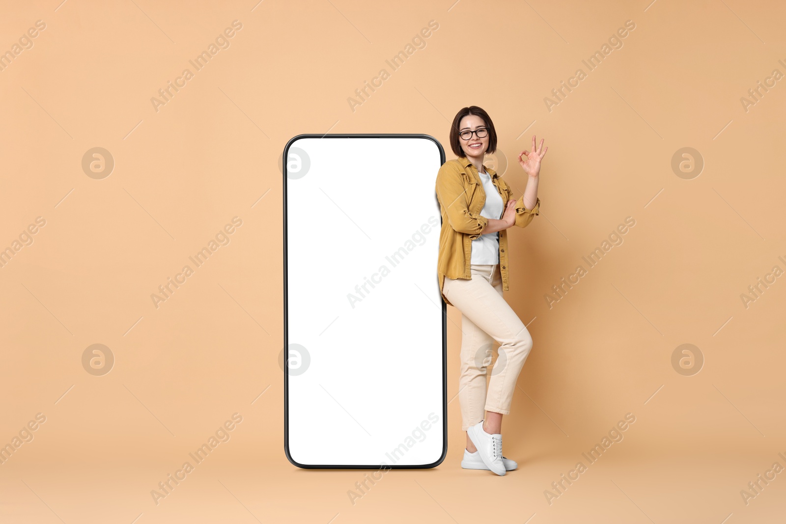 Image of Beautiful young woman showing Ok sign near big mobile phone with blank screen on dark beige background