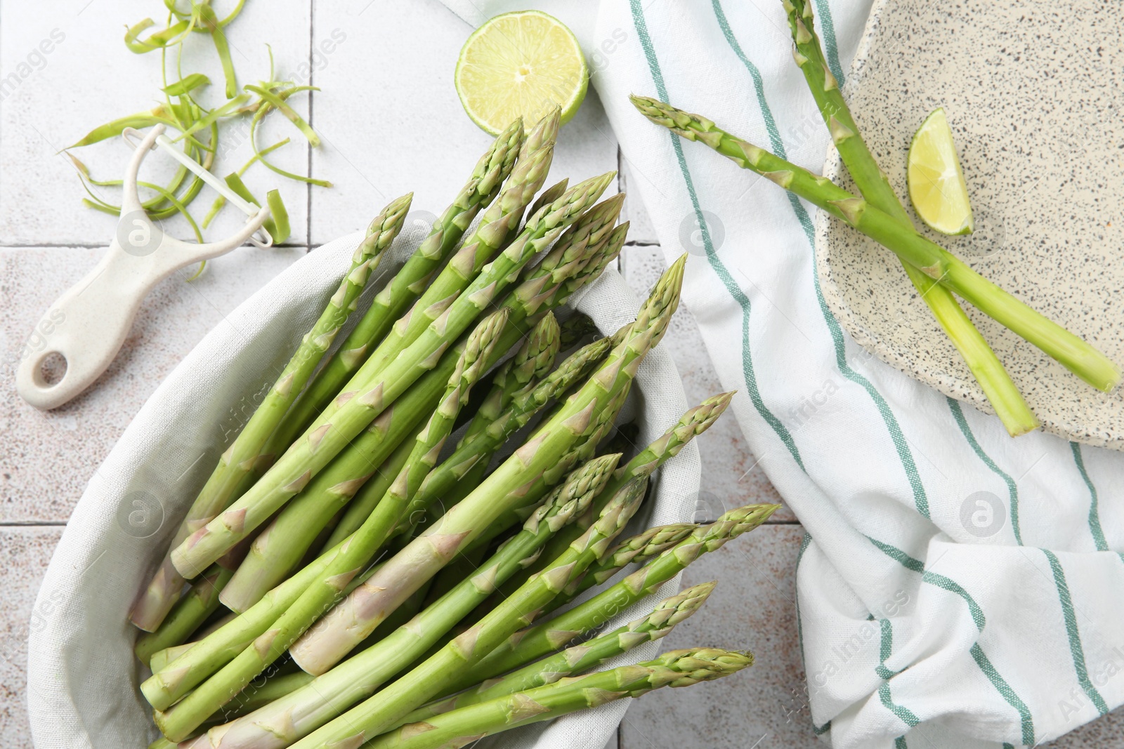 Photo of Fresh green asparagus stems, lime and vegetable peeler on light textured tiled table, flat lay