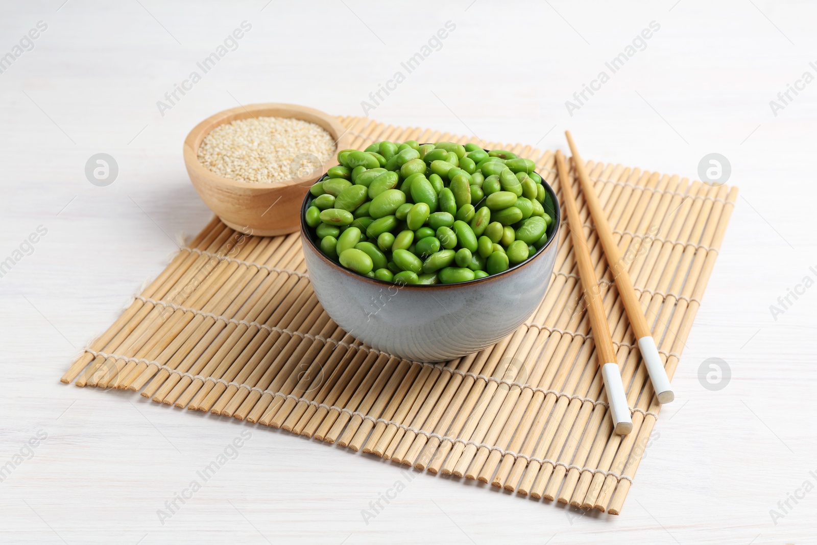 Photo of Fresh edamame soybeans in bowl, chopsticks and sesame seeds on white wooden table