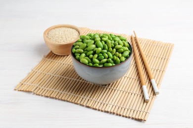 Fresh edamame soybeans in bowl, chopsticks and sesame seeds on white wooden table