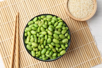 Photo of Fresh edamame soybeans in bowl, chopsticks and sesame seeds on white wooden table, top view