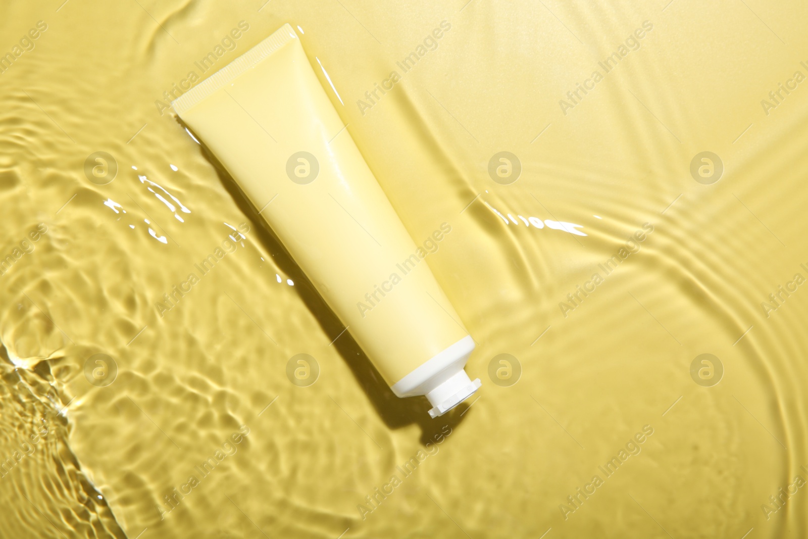 Photo of Tube with cream in water on yellow background, top view. Cosmetic product