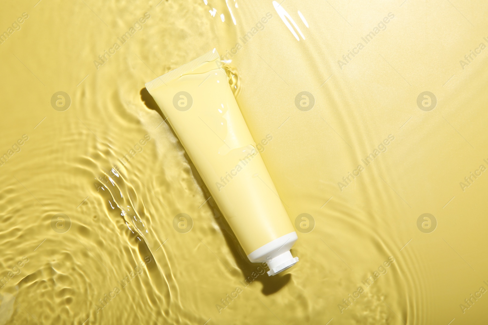 Photo of Tube with cream in water on yellow background, top view. Cosmetic product