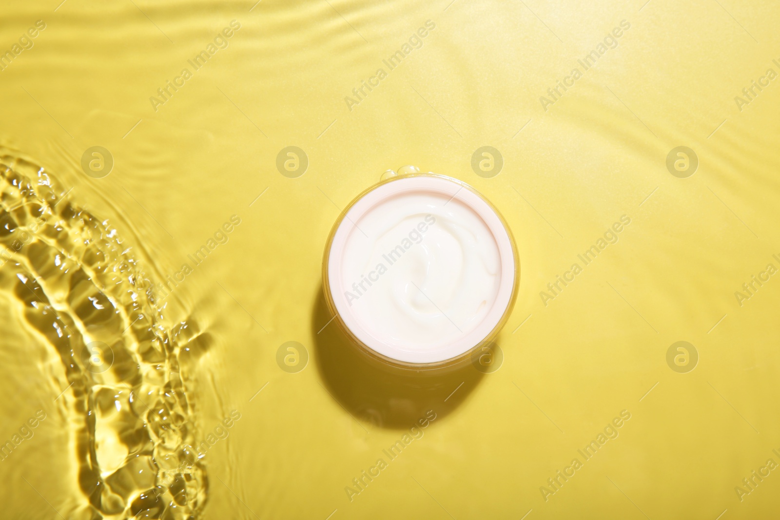 Photo of Jar with cream in water on yellow background, top view. Cosmetic product