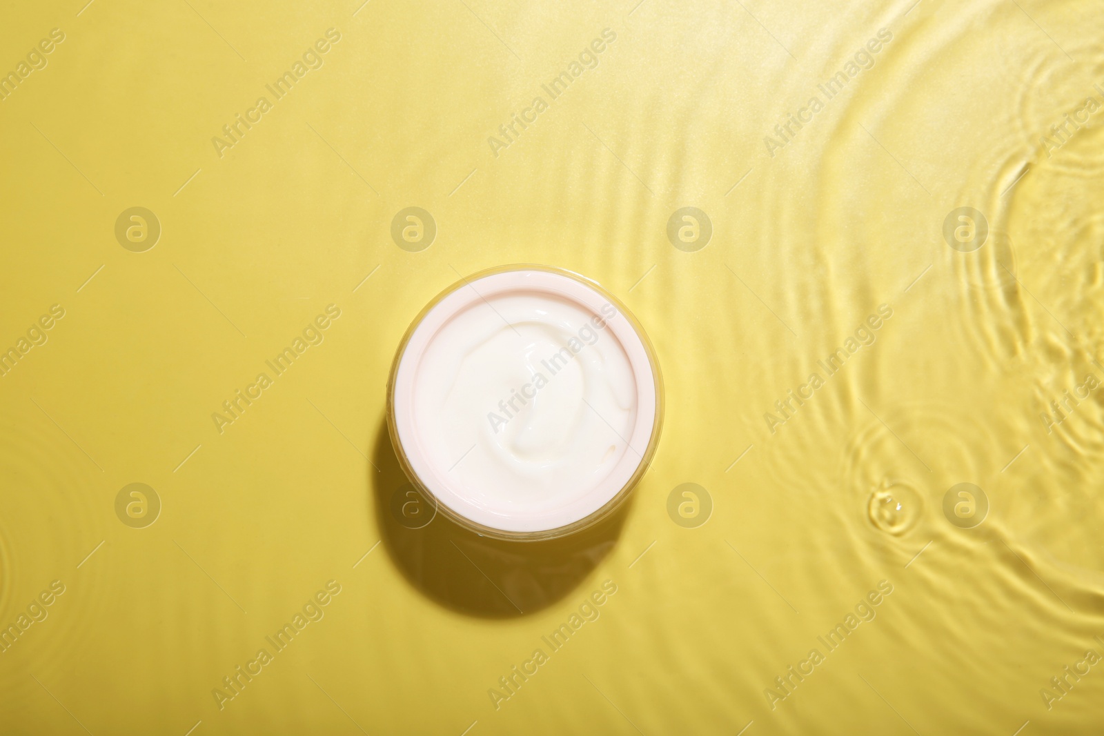 Photo of Jar with cream in water on yellow background, top view. Cosmetic product