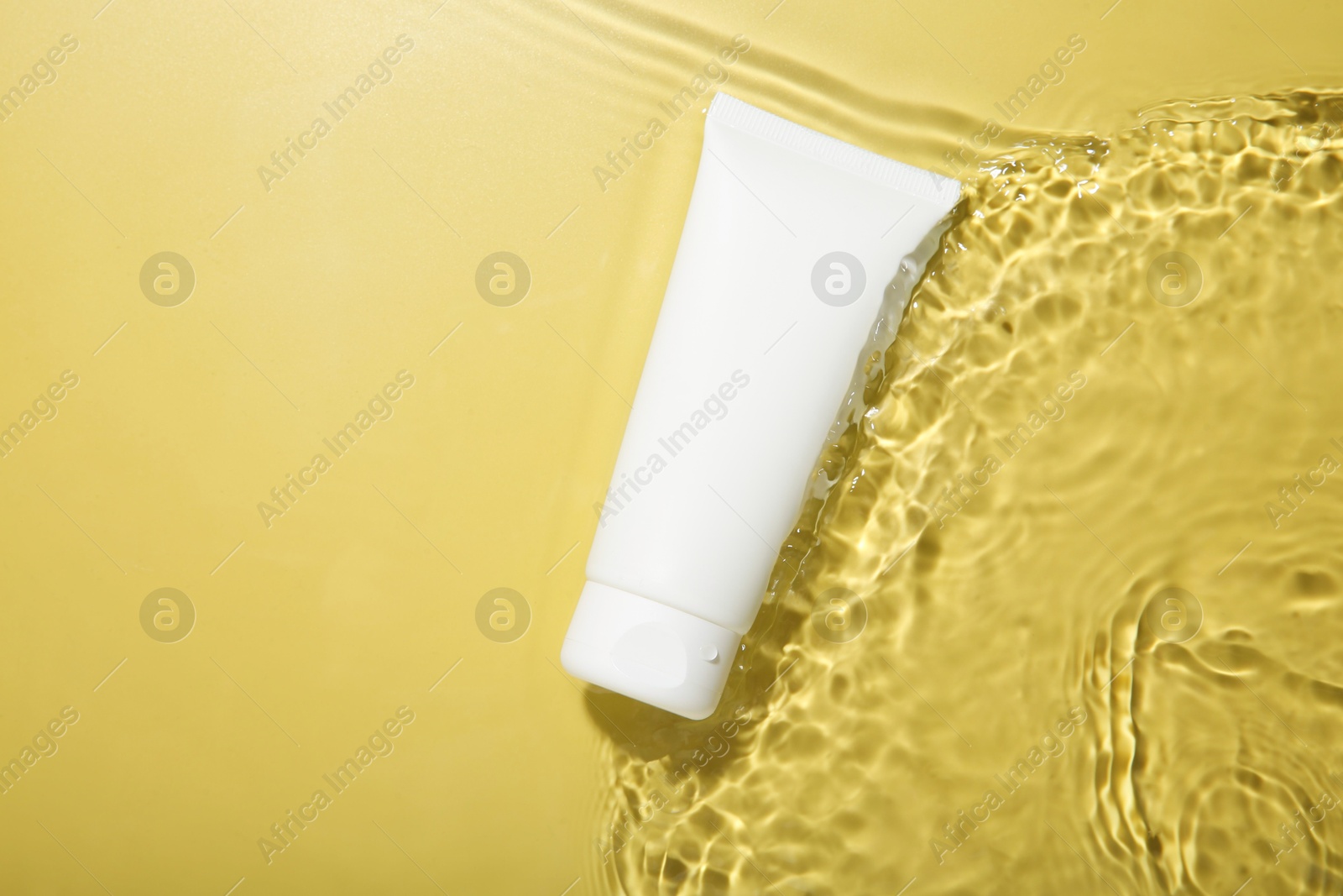 Photo of Tube with cream in water on yellow background, top view. Cosmetic product