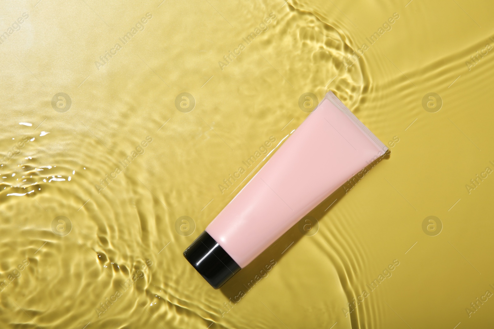 Photo of Tube with cream in water on yellow background, top view. Cosmetic product