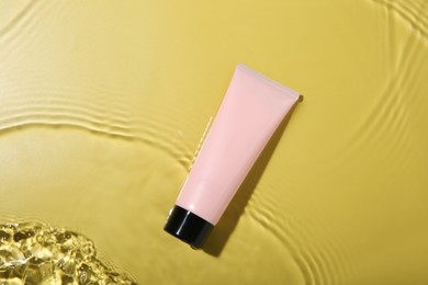 Photo of Tube with cream in water on yellow background, top view. Cosmetic product