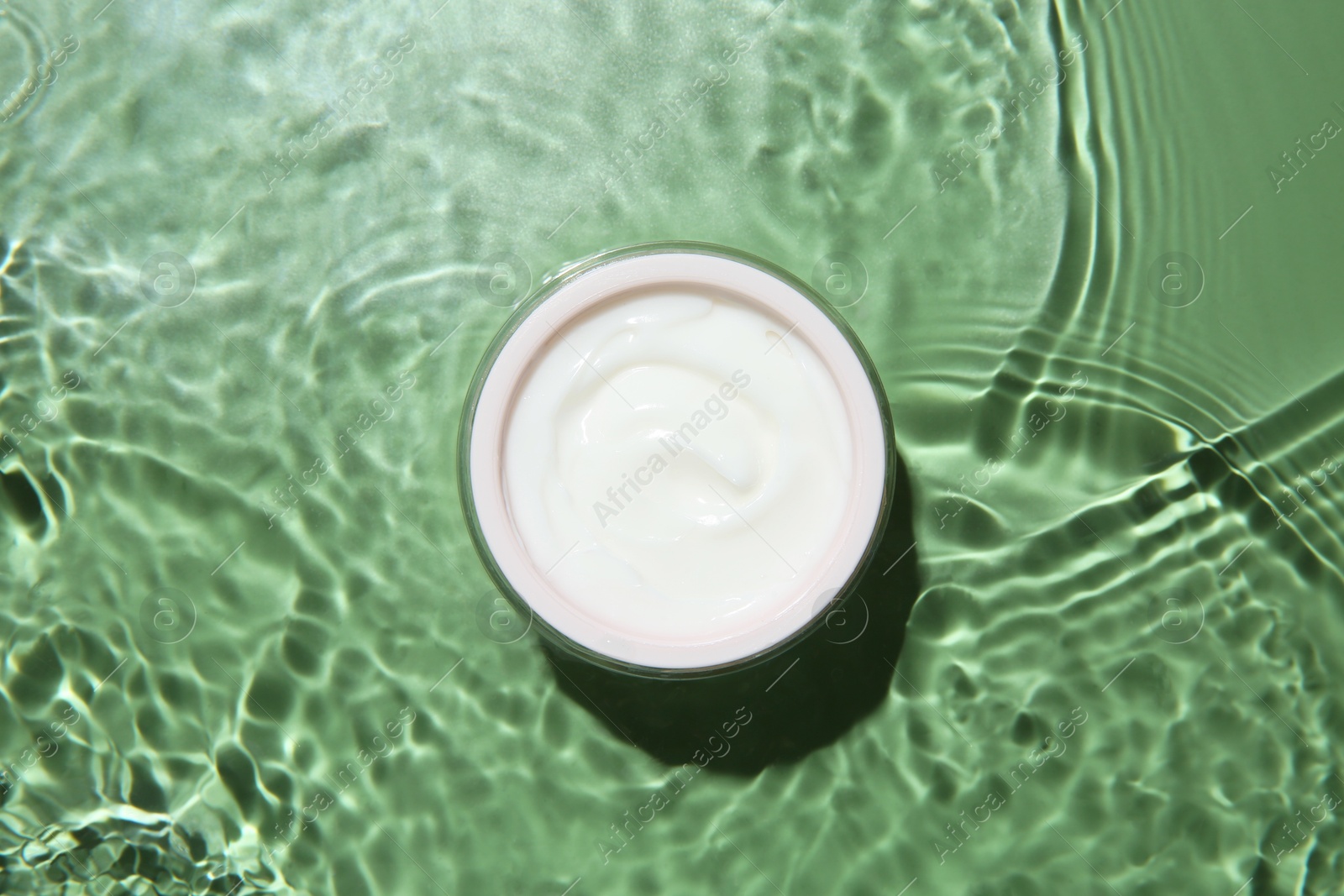 Photo of Jar with cream in water on light green background, top view. Cosmetic product