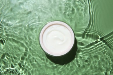 Photo of Jar with cream in water on light green background, top view. Cosmetic product