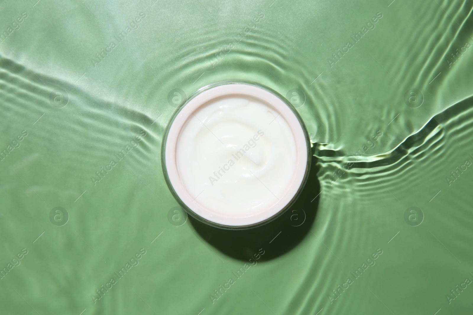 Photo of Jar with cream in water on light green background, top view. Cosmetic product