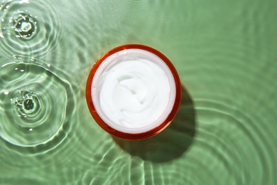 Photo of Jar with cream in water on light green background, top view. Cosmetic product