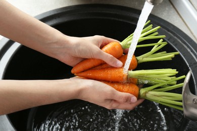 Woman washing fresh carrots under tap water in above sink indoors, closeup