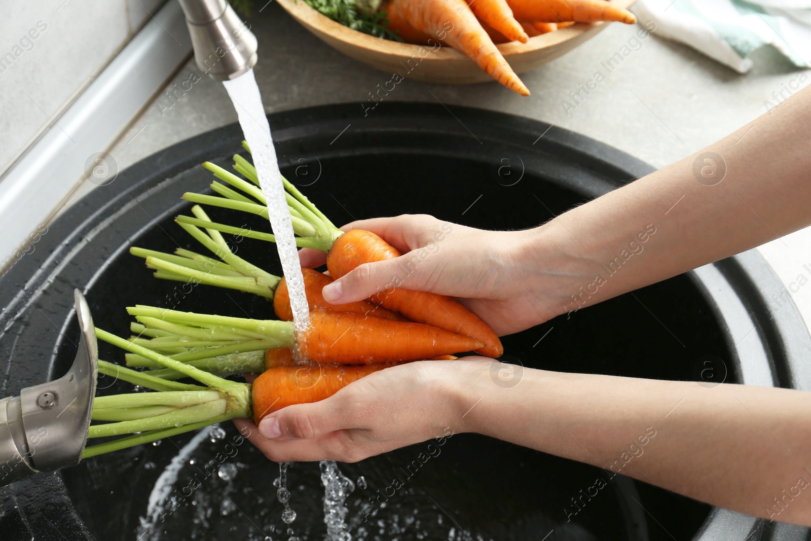 Photo of Woman washing fresh carrots under tap water in above sink indoors, closeup