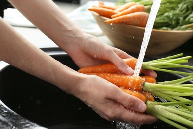 Photo of Woman washing fresh carrots under tap water in above sink indoors, closeup