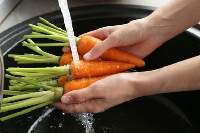 Photo of Woman washing fresh carrots under tap water in above sink indoors, closeup