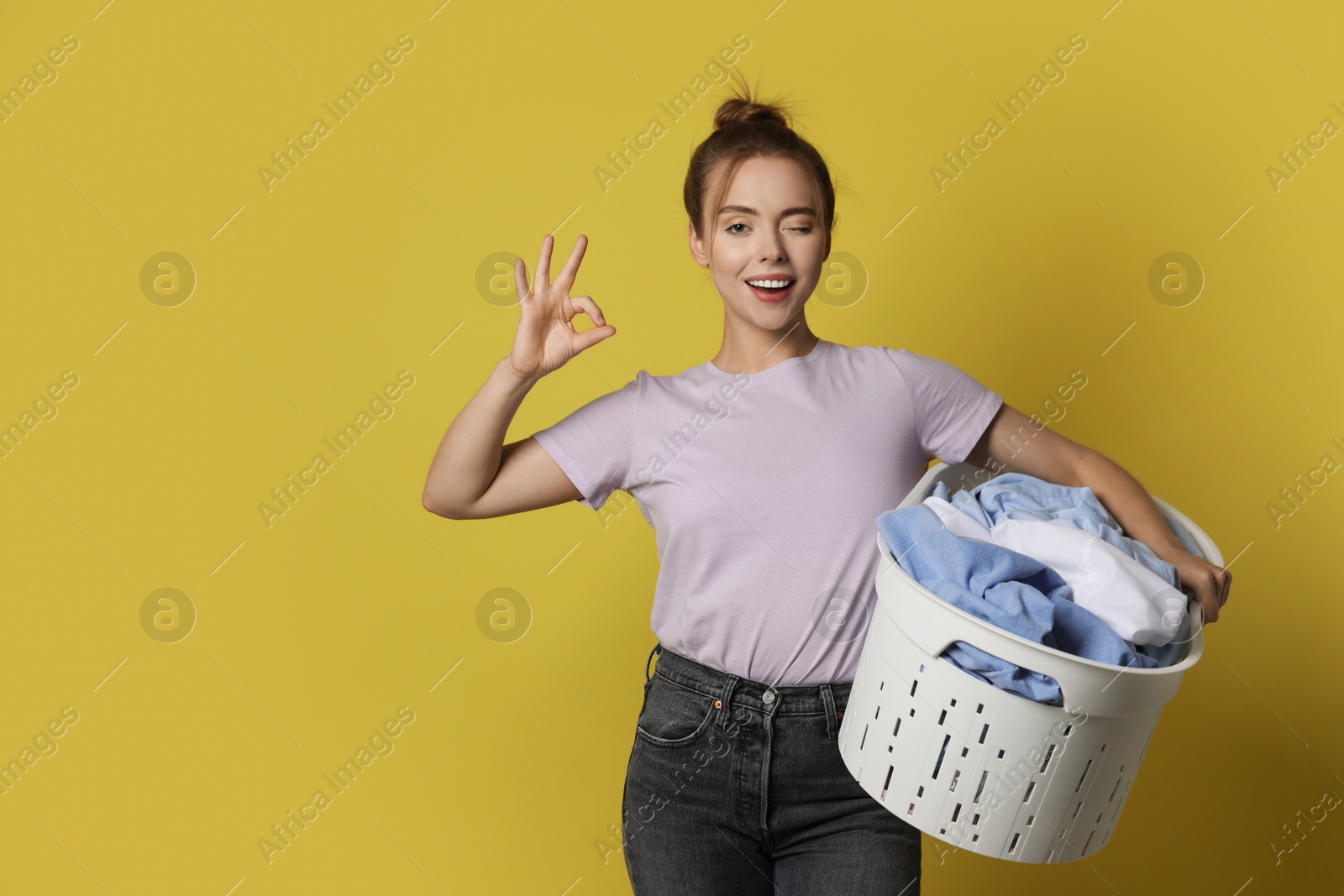 Photo of Happy young housewife with basket full of laundry showing OK gesture on yellow background. Space for text