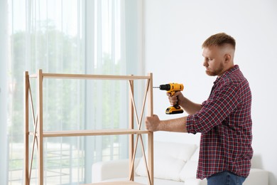 Photo of Man with electric screwdriver assembling wooden shelving unit in room