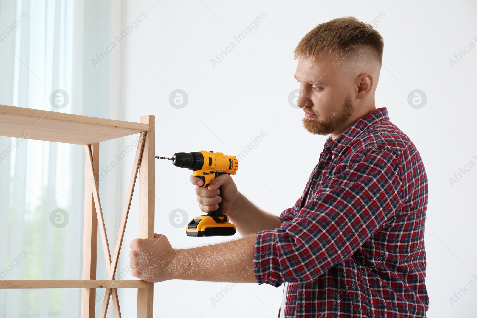 Photo of Man with electric screwdriver assembling wooden shelving unit in room