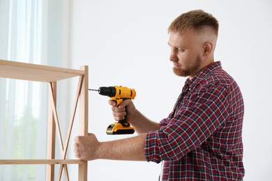 Photo of Man with electric screwdriver assembling wooden shelving unit in room