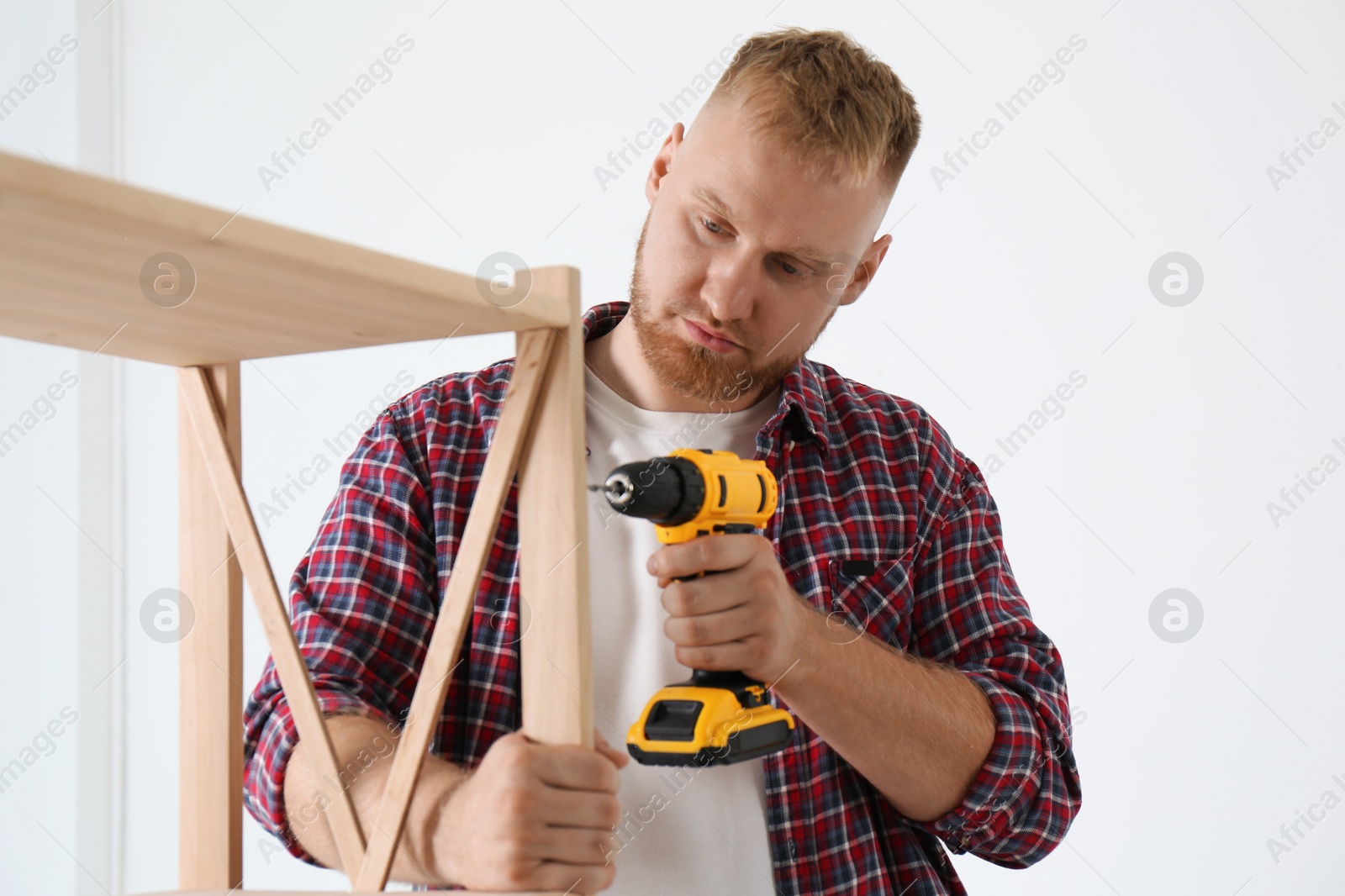 Photo of Man with electric screwdriver assembling wooden shelving unit at home