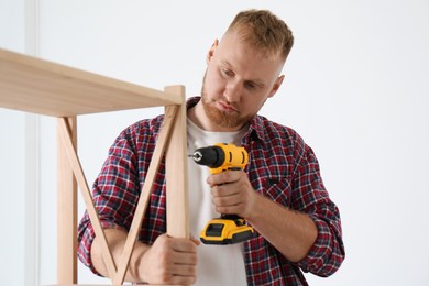 Photo of Man with electric screwdriver assembling wooden shelving unit at home