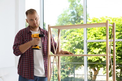 Photo of Man with electric screwdriver assembling wooden shelving unit at home