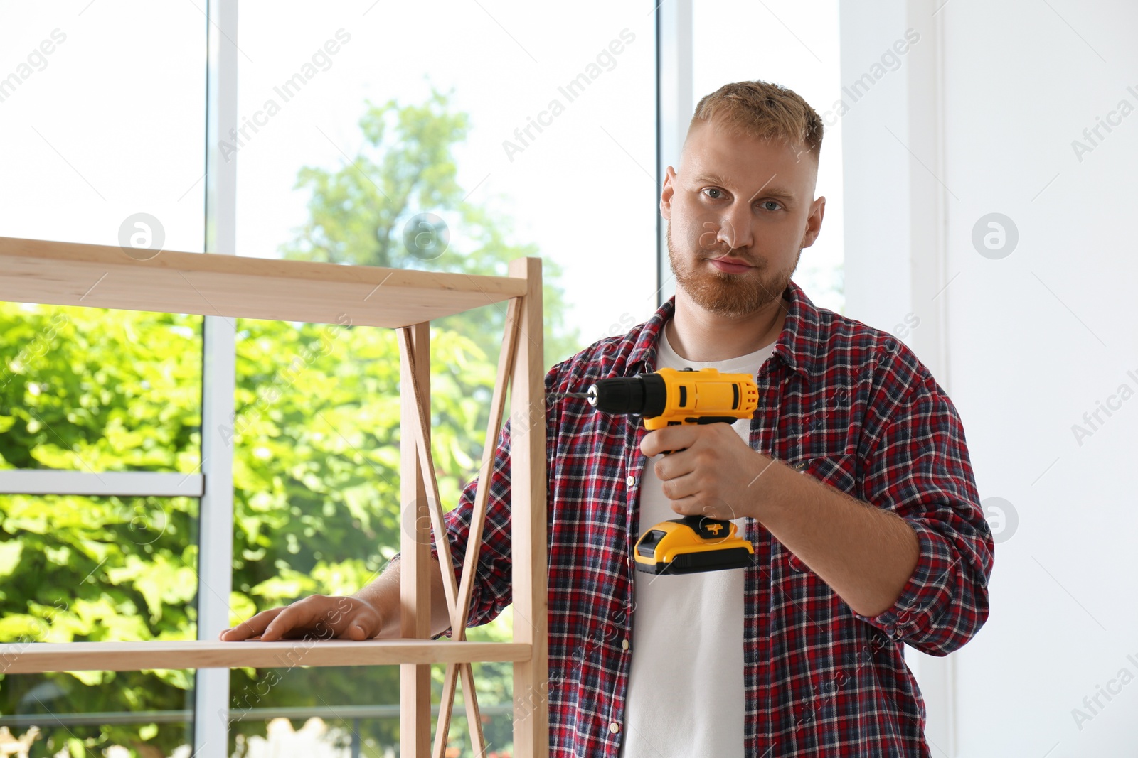 Photo of Man with electric screwdriver assembling wooden shelving unit at home