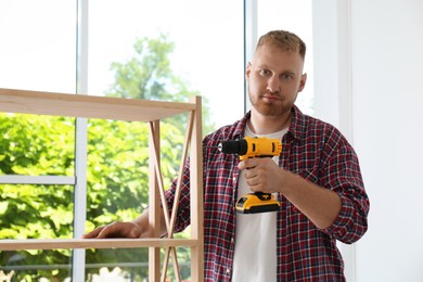 Photo of Man with electric screwdriver assembling wooden shelving unit at home