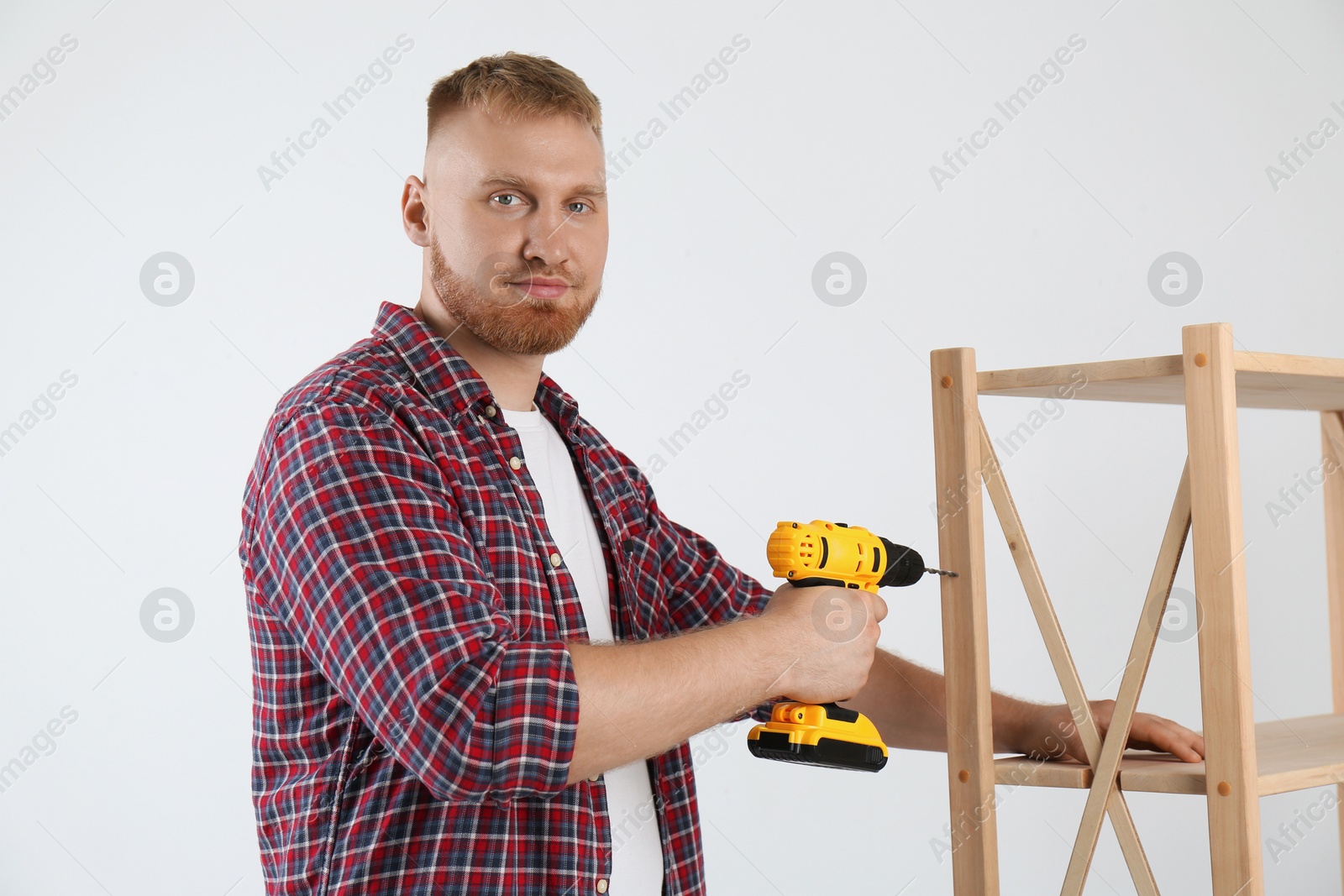 Photo of Man with electric screwdriver assembling furniture near white wall indoors