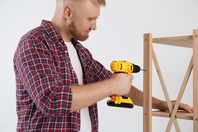 Photo of Man with electric screwdriver assembling furniture near white wall indoors