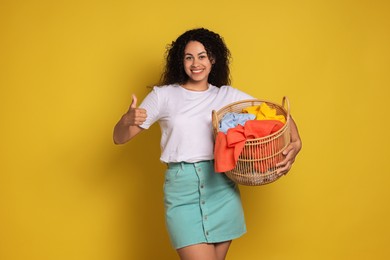 Photo of Happy woman with basket full of laundry showing thumbs up on yellow background