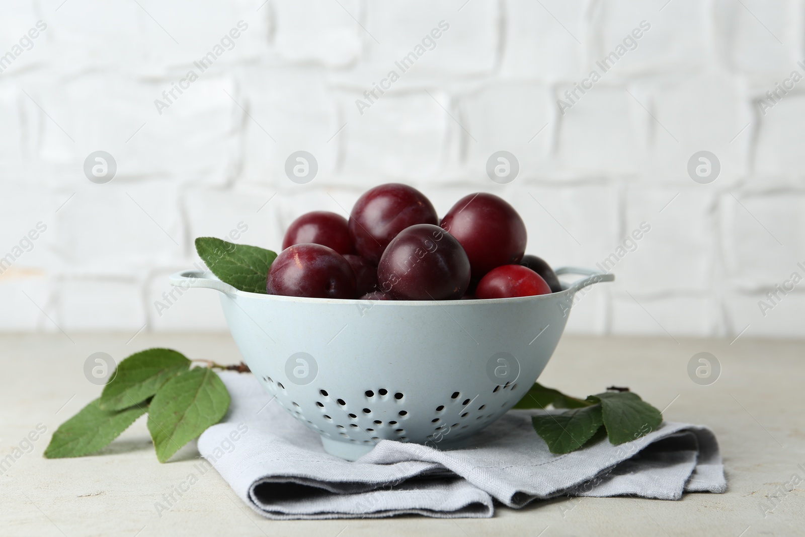 Photo of Ripe plums in colander and leaves on light textured table