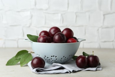 Photo of Ripe plums in colander and leaves on light textured table