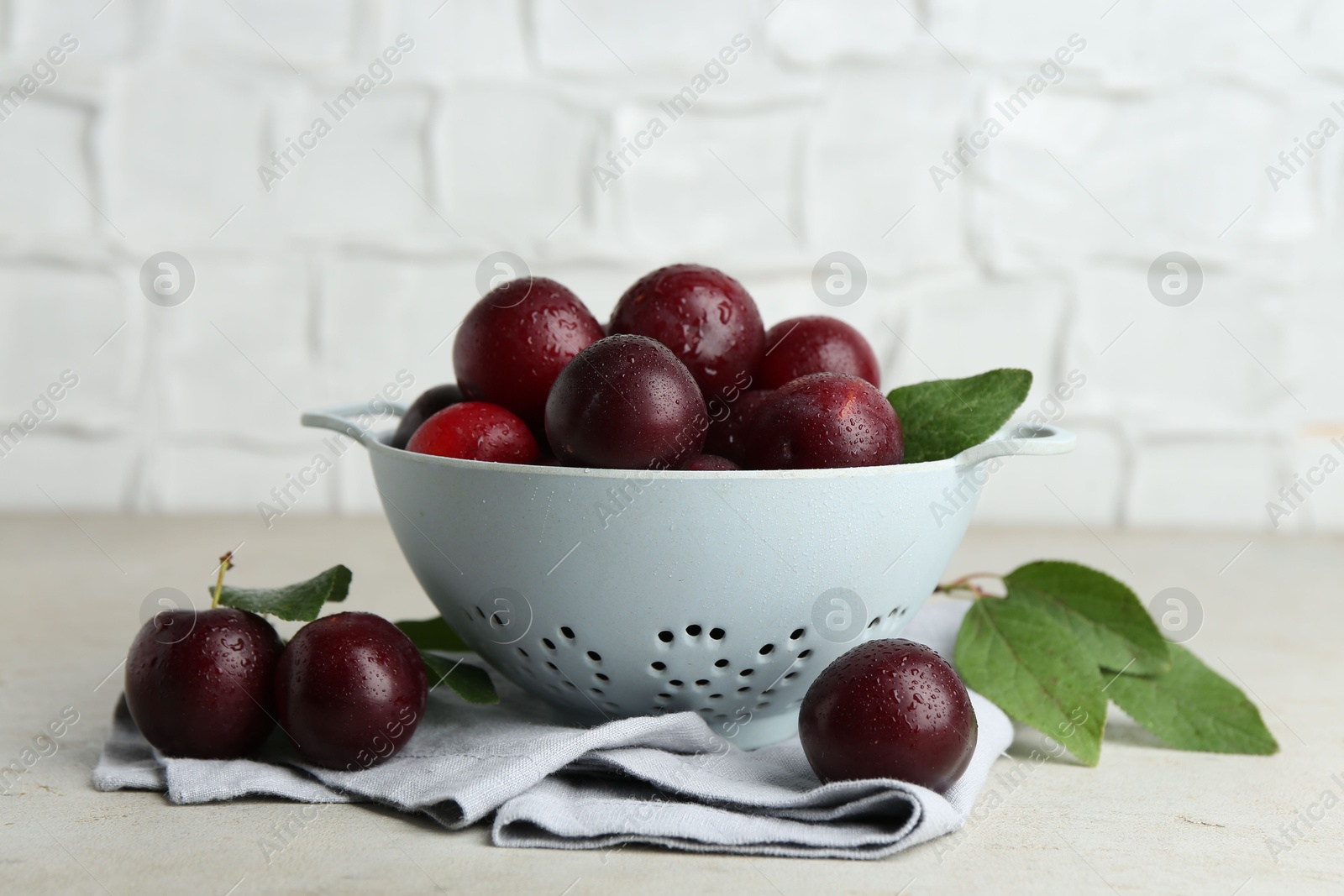 Photo of Ripe plums in colander and leaves on light textured table