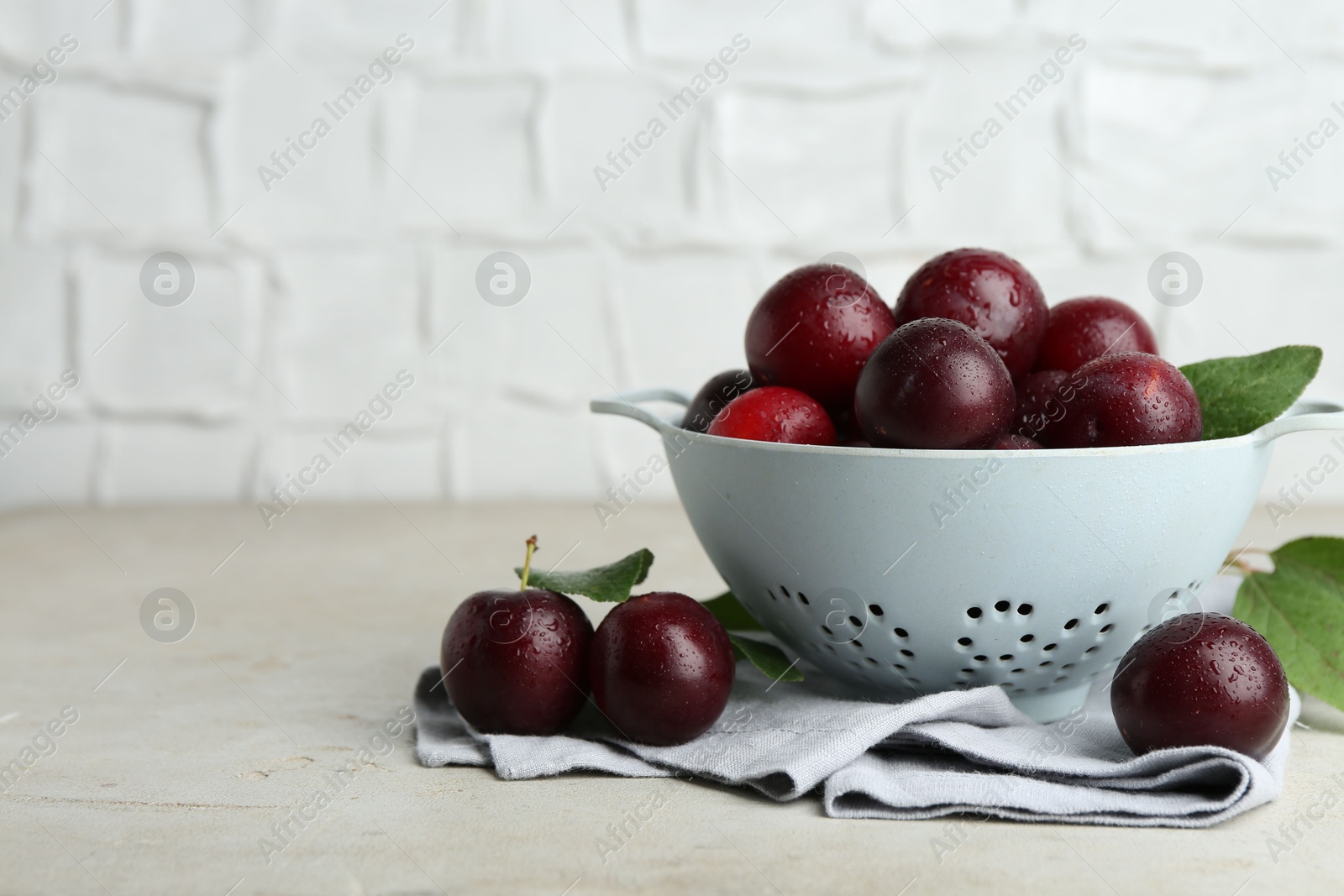 Photo of Ripe plums in colander and leaves on light textured table, space for text