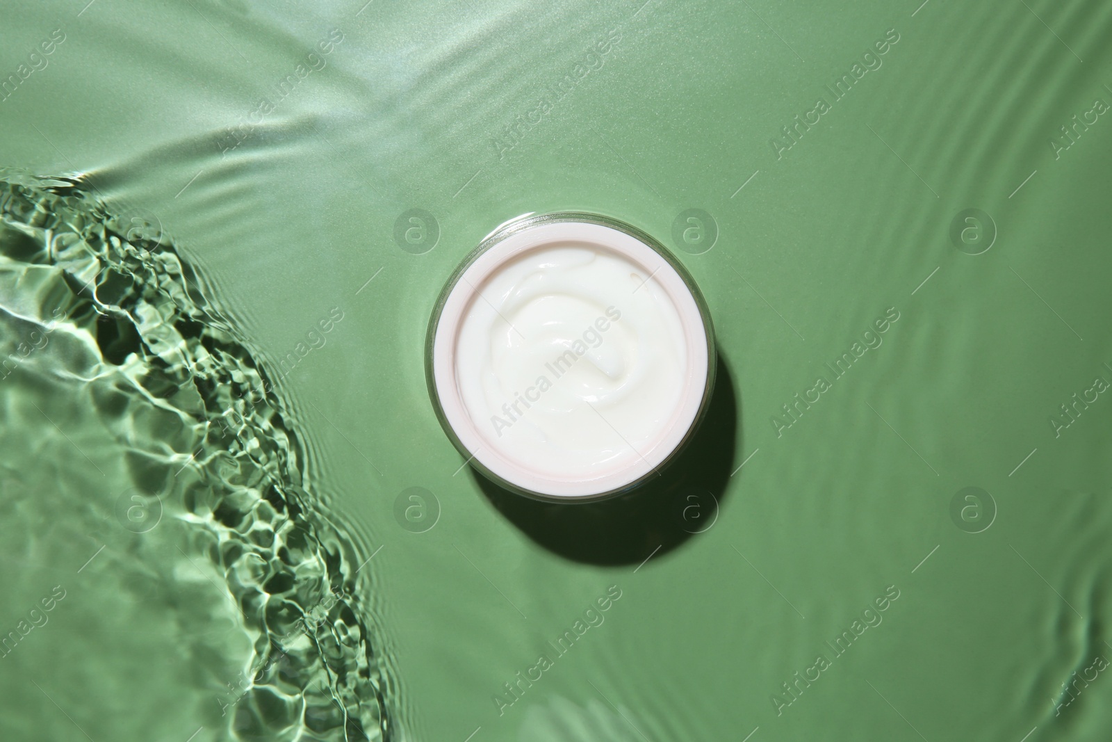 Photo of Jar with cream in water on light green background, top view. Cosmetic product