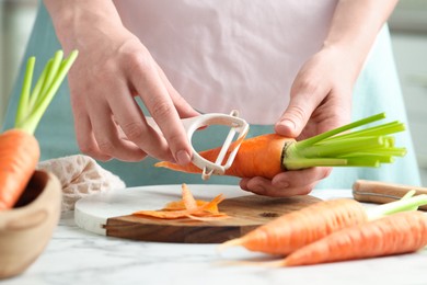 Woman peeling fresh carrot at white marble table, closeup