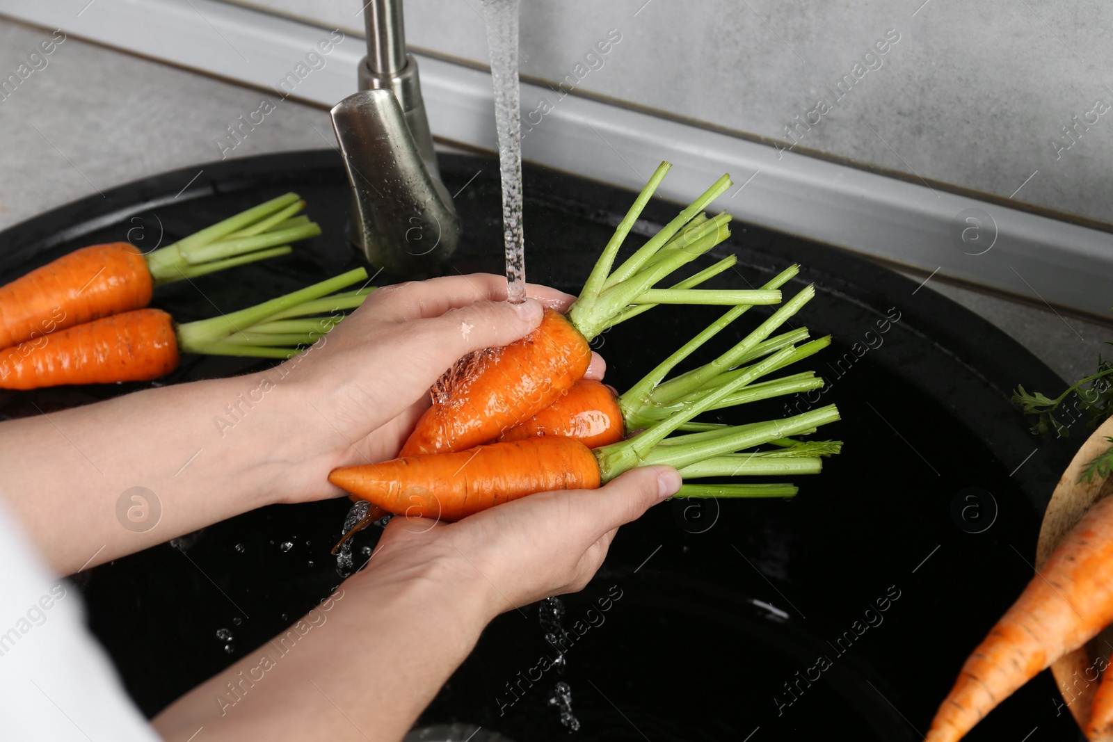 Photo of Woman washing fresh carrots under tap water in above sink indoors, closeup