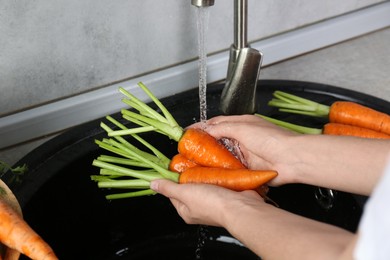 Woman washing fresh carrots under tap water in above sink indoors, closeup