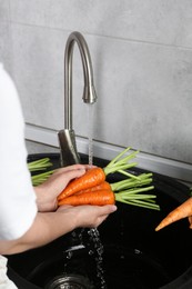 Photo of Woman washing fresh carrots under tap water in above sink indoors, closeup