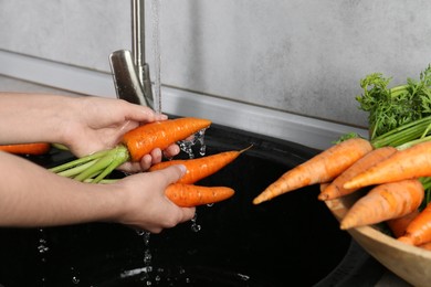 Woman washing fresh carrots under tap water in above sink indoors, closeup