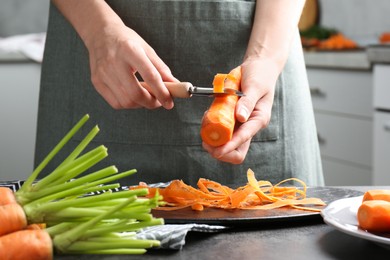 Woman peeling fresh carrot at dark gray table in kitchen, closeup