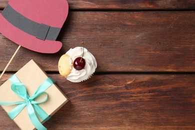 Photo of Happy Father's Day. Cupcake, paper hat and gift box on wooden table, flat lay. Space for text