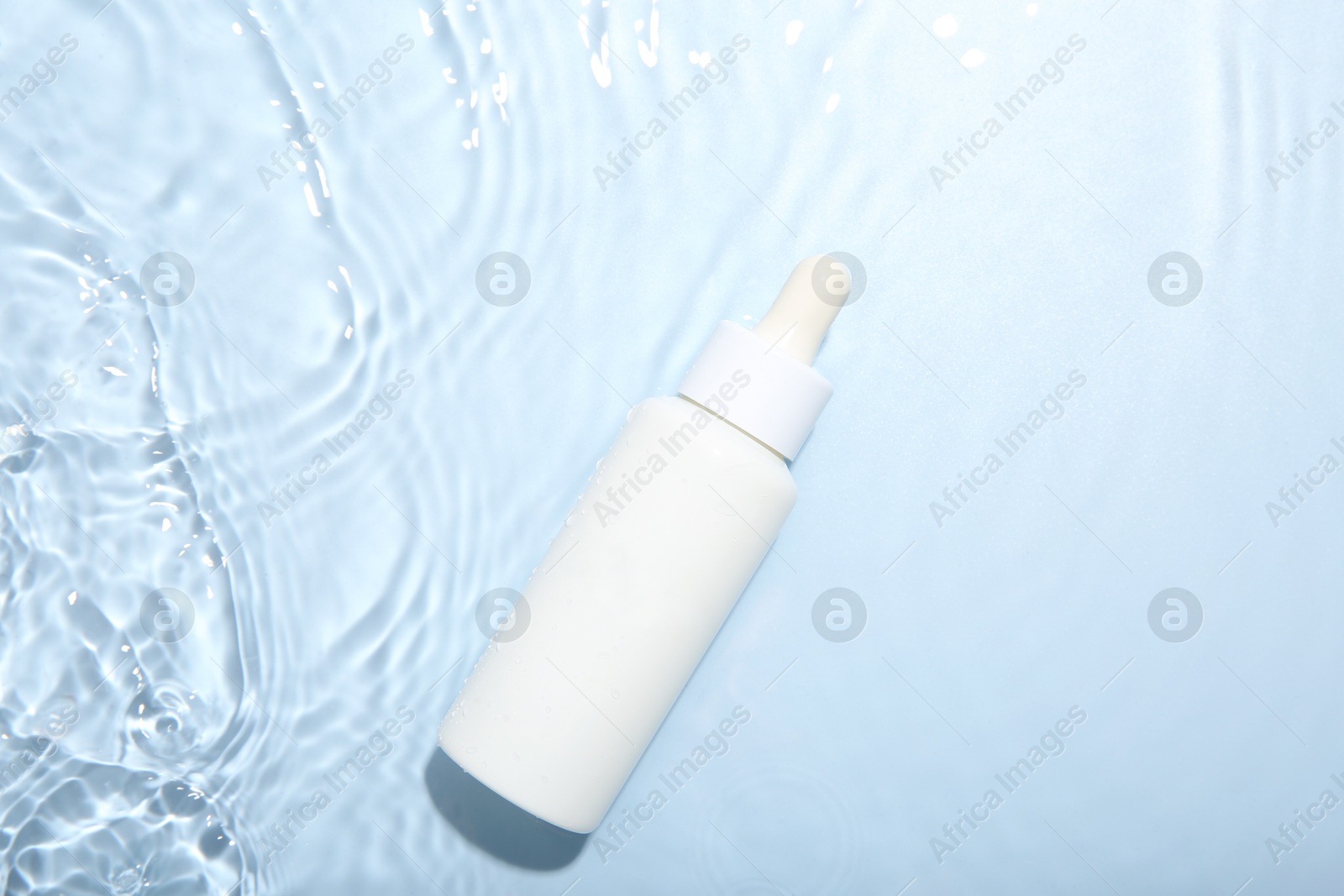 Photo of Bottle of cosmetic product in water on light blue background, top view