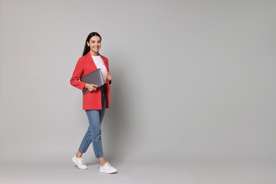 Beautiful woman in red jacket with laptop on gray background, space for text