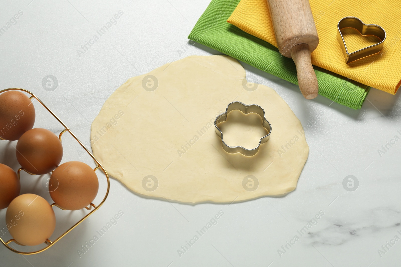 Photo of Raw dough, cookie cutters, eggs, napkins and rolling pin on white marble table, above view