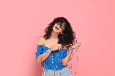 Happy young woman wearing carnival mask and tinsel on pink background